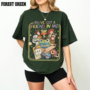 May include: A forest green short-sleeve t-shirt featuring a colorful graphic print of Toy Story characters. The design includes the text "You've Got A Friend In Me" and "Toy Story 5" with a central box labeled "Jason" surrounded by various animated figures.