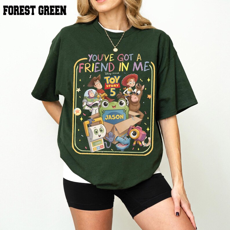 May include: A forest green short-sleeve t-shirt featuring a colorful graphic print of Toy Story characters. The design includes the text "You've Got A Friend In Me" and "Toy Story 5" with a central box labeled "Jason" surrounded by various animated figures.
