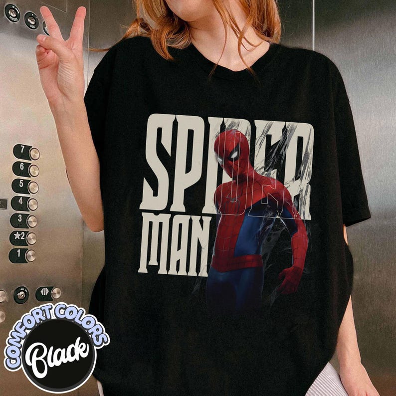Peut inclure: T-shirt noir avec l'inscription "SPIDER-MAN" en gras, en blanc cass&eacute;, et une image de Spider-Man en costume rouge et bleu. Le t-shirt est de la marque Comfort Colors.