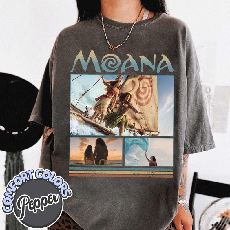 May include: A charcoal grey short-sleeve t-shirt featuring a vintage-style graphic print of the movie Moana. The design includes the text MOANA, a scene of characters on a sailboat, a sunset beach silhouette, and a character reaching toward blue water.