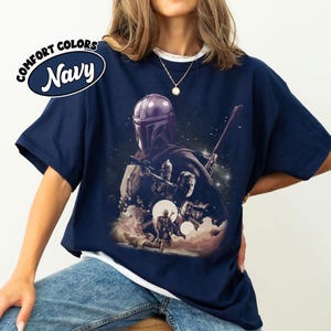 K&ouml;nnte beinhalten: Marineblaues T-Shirt mit einer Grafik mit Figuren aus einem Science-Fiction-Film. Das Shirt hat die Worte "COMFORT COLORS" und "Navy" in einem kreisf&ouml;rmigen Design. Die Grafik zeigt eine Figur mit Helm und andere Charaktere.