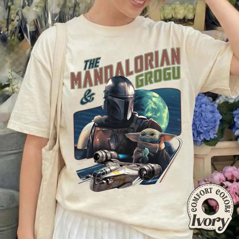 May include: Ivory-colored t-shirt featuring a graphic of The Mandalorian and Grogu, with a spaceship and planet in the background. The text "The Mandalorian & Grogu" is printed above the graphic in brown and green.