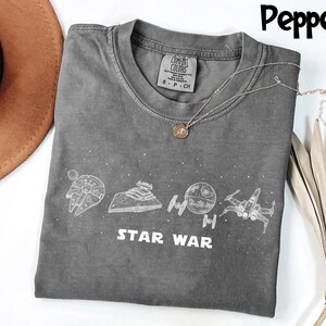 May include: A gray Comfort Colors t-shirt featuring white line art of Star Wars spacecraft and the words "STAR WAR". A silver necklace with a pendant rests on the shirt. The shirt is folded.
