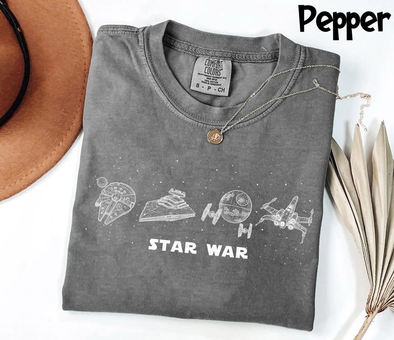 May include: A gray Comfort Colors t-shirt featuring white line art of Star Wars spacecraft and the words "STAR WAR". A silver necklace with a pendant rests on the shirt. The shirt is folded.