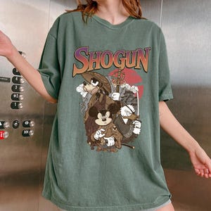 Puede incluir: Una camiseta de manga corta color verde salvia con un estampado gr&aacute;fico que incluye el texto SHOGUN en una fuente degradada. La ilustraci&oacute;n muestra a tres personajes de dibujos animados vestidos con atuendos japoneses tradicionales.
