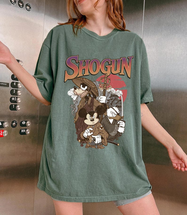 Puede incluir: Una camiseta de manga corta color verde salvia con un estampado gr&aacute;fico que incluye el texto SHOGUN en una fuente degradada. La ilustraci&oacute;n muestra a tres personajes de dibujos animados vestidos con atuendos japoneses tradicionales.
