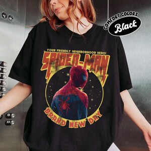 May include: Black t-shirt featuring a graphic of Spider-Man in a yellow circle with the text "YOUR FRIENDLY NEIGHBORHOOD HERO! SPIDER-MAN BRAND NEW DAY". The shirt is labeled "COMFORT COLORS Black".