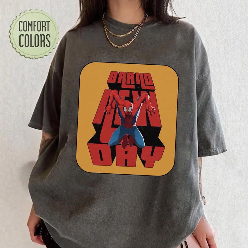 Puede incluir: Camiseta gris oscuro con un gr&aacute;fico de Spider-Man y las palabras "BRAND NEW DAY" en rojo y amarillo. La camiseta tiene un aspecto vintage, y las palabras "COMFORT COLORS" est&aacute;n en un c&iacute;rculo verde.