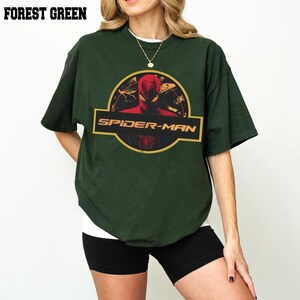 May include: A forest green short-sleeved t-shirt featuring a circular graphic with a red Spider-Man character illustration and the text SPIDER-MAN in a yellow font. The design is centred on the chest of the garment, which has a crew neckline.