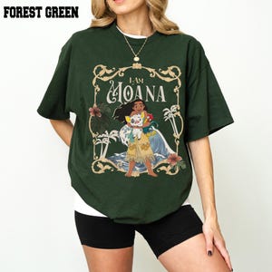 May include: A forest green short-sleeved t-shirt featuring a graphic print of the character Moana holding a pig and a rooster. The design is enclosed in an ornate gold-colored frame with tropical foliage and the text I AM MOANA.