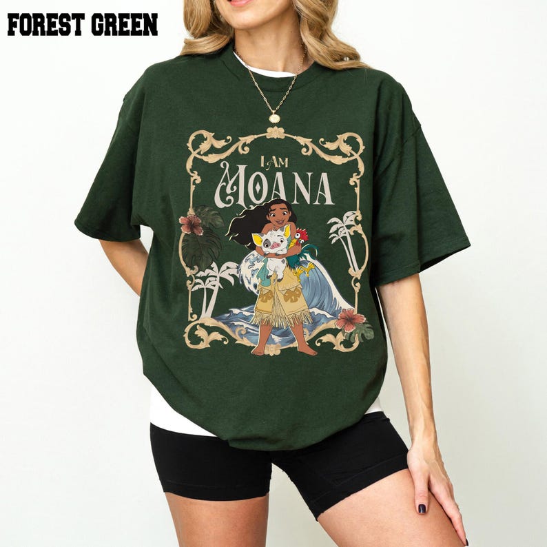 May include: A forest green short-sleeved t-shirt featuring a graphic print of the character Moana holding a pig and a rooster. The design is enclosed in an ornate gold-colored frame with tropical foliage and the text I AM MOANA.