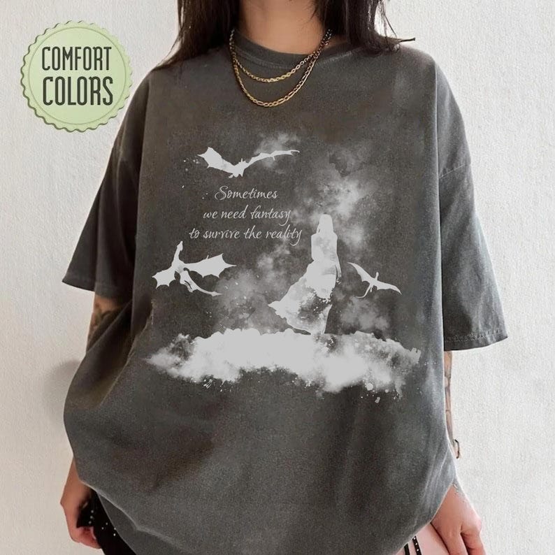 May include: A charcoal gray short-sleeve t-shirt featuring a white graphic print of a silhouette standing amidst clouds with two flying dragons. The text reads: Sometimes we need fantasy to survive the reality. A Comfort Colors logo is in the top left.