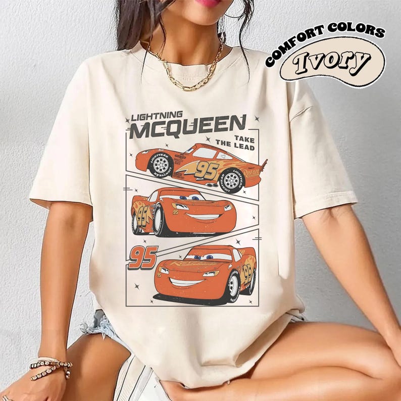 May include: Ivory-colored t-shirt featuring a graphic of Lightning McQueen from the movie Cars. The design includes three images of the character with the number 95, and the text "Lightning McQueen" and "Take the Lead."