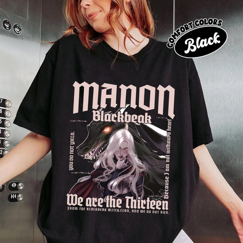 May include: Black t-shirt featuring the text "MANON Blackbeak" in pink, with a graphic of a woman with white hair and a dragon. The shirt also includes the phrases "You do not yield" and "We are the Thirteen."