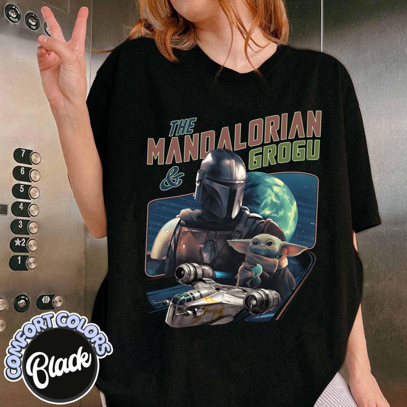 May include: Black t-shirt featuring a graphic of The Mandalorian and Grogu, with the text "The Mandalorian & Grogu." The design includes a spaceship and a planet in the background. The shirt is labeled "Comfort Colors Black."