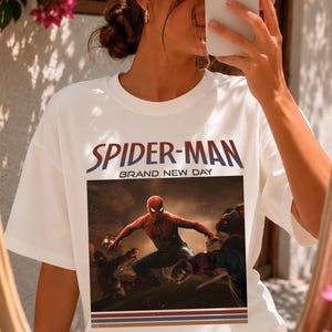 May include: A white short-sleeve t-shirt featuring a graphic print of Spider-Man in a dynamic pose. The text "SPIDER-MAN" appears in large letters above the image, with "BRAND NEW DAY" underneath. The design includes three horizontal stripes at the bottom.