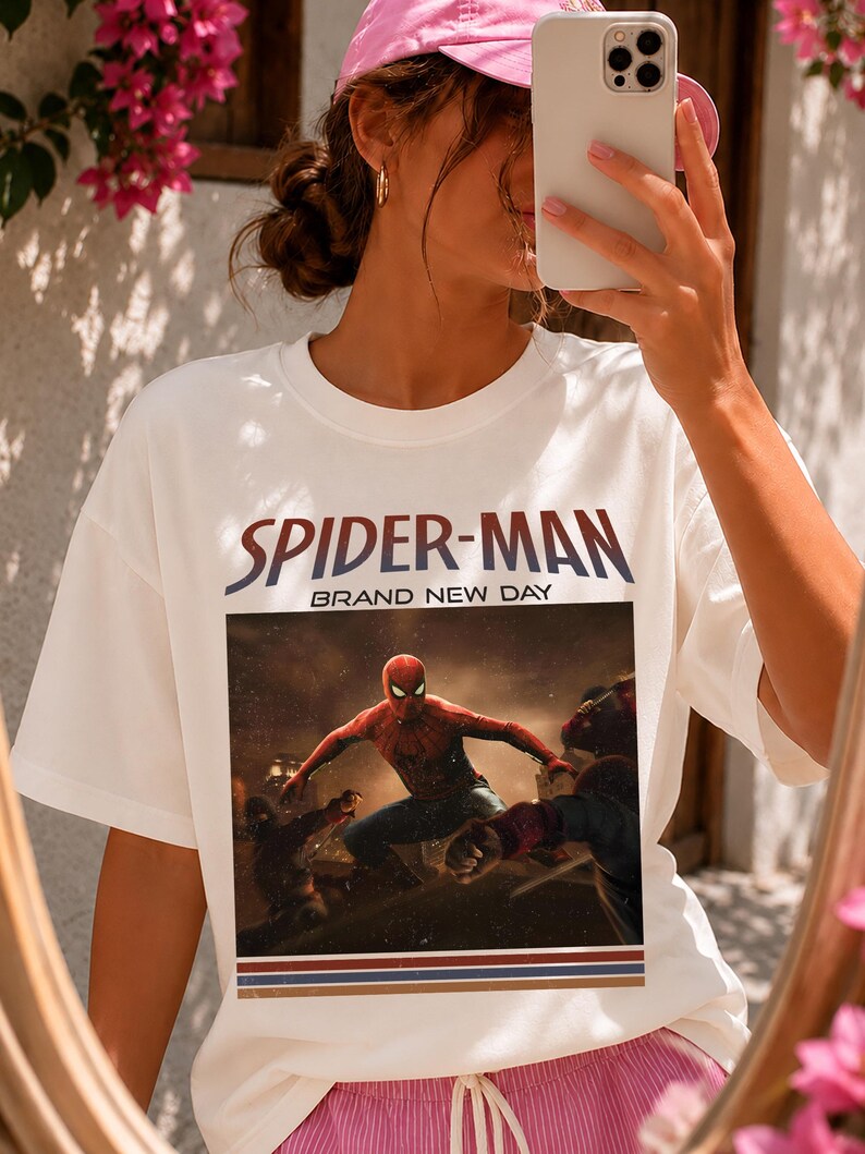 May include: A white short-sleeve t-shirt featuring a graphic print of Spider-Man in a dynamic pose. The text "SPIDER-MAN" appears in large letters above the image, with "BRAND NEW DAY" underneath. The design includes three horizontal stripes at the bottom.