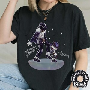 Pu&ograve; includere: Una t-shirt nera con un'illustrazione grafica di Michael Jackson e Mickey Mouse che ballano su una piattaforma luminosa. Il design include accenti a forma di stella bianca e il testo Michael Jackson e Mickey Mouse in carattere corsivo.