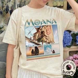 May include: An ivory short-sleeve t-shirt featuring the text MOANA in blue and a graphic collage of scenes from the movie Moana, including characters on a boat, a sunset silhouette, and a person holding a blue cloth, with striped accents.