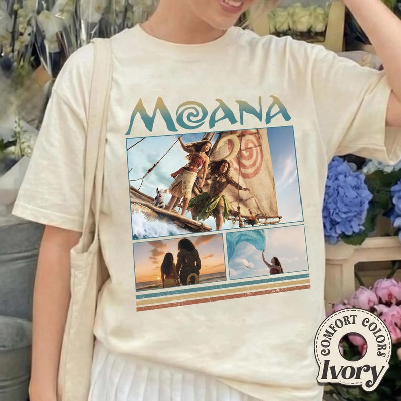 May include: An ivory short-sleeve t-shirt featuring the text MOANA in blue and a graphic collage of scenes from the movie Moana, including characters on a boat, a sunset silhouette, and a person holding a blue cloth, with striped accents.