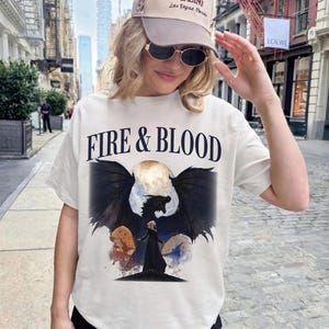 May include: A cream-colored short-sleeve t-shirt featuring the text FIRE & BLOOD in black lettering above a graphic of a black dragon with spread wings and a figure in a black cloak standing before a large moon, paired with a beige baseball cap.