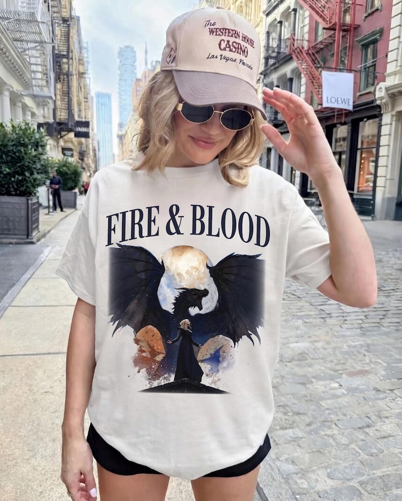 May include: A cream-colored short-sleeve t-shirt featuring the text FIRE & BLOOD in black lettering above a graphic of a black dragon with spread wings and a figure in a black cloak standing before a large moon, paired with a beige baseball cap.