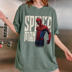 Peut inclure: T-shirt vert avec l'inscription "SPIDER MAN" en blanc, avec un effet vieilli. Le motif comprend une illustration de Spider-Man en rouge et bleu. Le t-shirt a des manches courtes et un col rond.