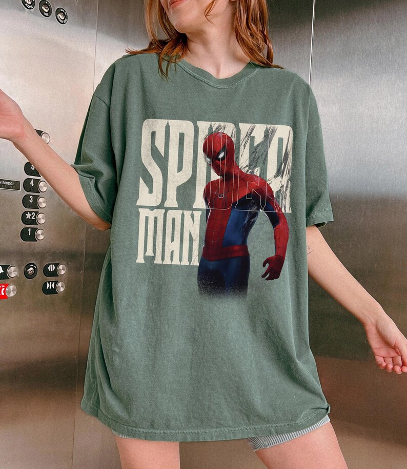 Peut inclure: T-shirt vert avec l'inscription "SPIDER MAN" en blanc, avec un effet vieilli. Le motif comprend une illustration de Spider-Man en rouge et bleu. Le t-shirt a des manches courtes et un col rond.