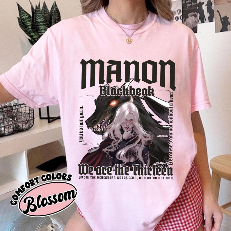 May include: A light pink t-shirt with the text "MANON Blackbeak" in black gothic font. Below is a graphic of a woman with white hair and a dragon. The text "We are the Thirteen" is also visible.