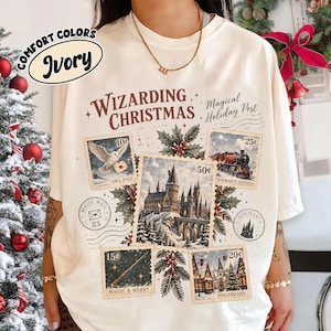 May include: An ivory t-shirt featuring a Wizarding Christmas graphic with multiple postage stamp illustrations. The design includes a snowy castle, an owl, a train, a wand, and a village, with text reading Wizarding Christmas and Magical Holiday Post.