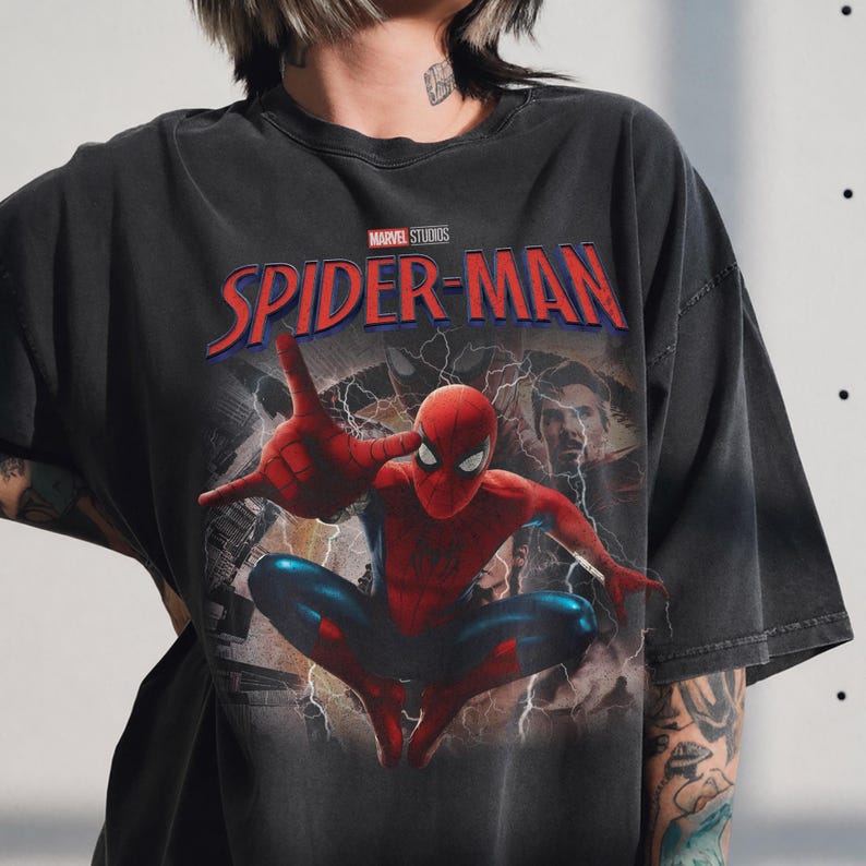 May include: A charcoal grey short-sleeve t-shirt featuring a graphic print of Spider-Man in a crouching pose with a web-shooting hand gesture. Above the character, the text reads Marvel Studios Spider-Man in red and blue lettering with lightning.