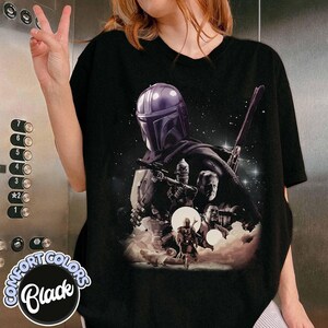 K&ouml;nnte beinhalten: Schwarzes T-Shirt mit einer Grafik eines Charakters mit Helm, der eine Waffe h&auml;lt, mit anderen Charakteren und einem Sternenhintergrund. Das Shirt hat die Worte "COMFORT COLORS" und "Black" in einem kreisf&ouml;rmigen Design.