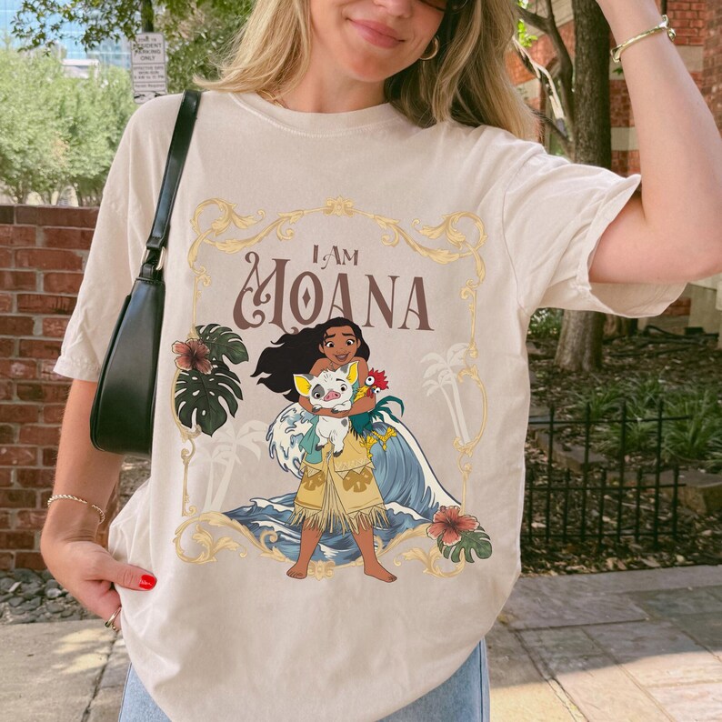 May include: A beige short-sleeve t-shirt featuring a graphic print of the character Moana holding a pig and a rooster. The design includes the text I AM MOANA, blue ocean waves, tropical leaves, and a gold ornate frame border around the illustration.