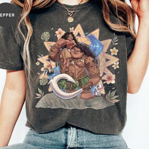 May include: A charcoal gray short-sleeve t-shirt featuring a graphic print of Moana and Maui from the Disney film. The design includes a large white fishhook, a rooster, a pig, and tropical flowers set against a yellow and orange sunburst background.