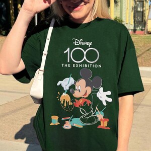 May include: Dark green t-shirt featuring the Disney 100 logo and Mickey Mouse as an artist. The graphic shows Mickey painting with paint pots and the text "THE EXHIBITION". The shirt is a short-sleeved, casual style.