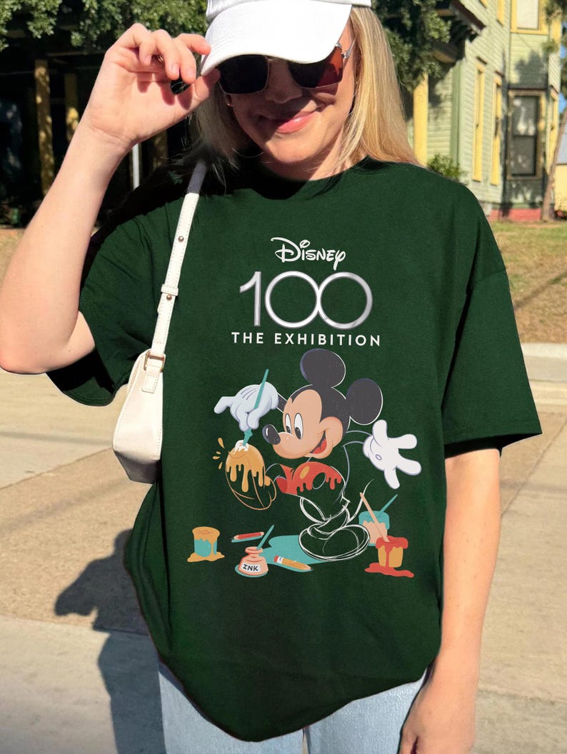 May include: Dark green t-shirt featuring the Disney 100 logo and Mickey Mouse as an artist. The graphic shows Mickey painting with paint pots and the text "THE EXHIBITION". The shirt is a short-sleeved, casual style.