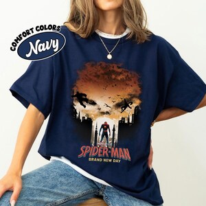 Może przedstawiać: Granatowa koszulka z grafiką Spider-Mana. Projekt zawiera sylwetki Spider-Mana w akcji, krajobraz miejski i napis "SPIDER-MAN BRAND NEW DAY". Koszulka ma r&oacute;wnież napis "COMFORT COLORS NAVY".