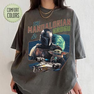 May include: Dark gray t-shirt featuring a graphic of The Mandalorian and Grogu, with a spaceship and planet in the background. The text "The Mandalorian & Grogu" is printed above the image in red and green.