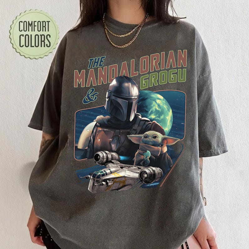 May include: Dark gray t-shirt featuring a graphic of The Mandalorian and Grogu, with a spaceship and planet in the background. The text "The Mandalorian & Grogu" is printed above the image in red and green.