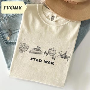 May include: Ivory-colored t-shirt featuring a Star Wars design. The graphic includes illustrations of the Millennium Falcon, a Star Destroyer, the Death Star, an X-wing fighter, and the words "STAR WAR" in black.