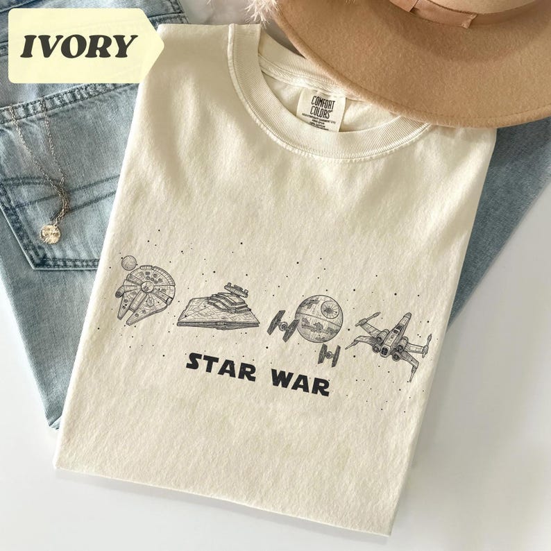 May include: Ivory-colored t-shirt featuring a Star Wars design. The graphic includes illustrations of the Millennium Falcon, a Star Destroyer, the Death Star, an X-wing fighter, and the words "STAR WAR" in black.
