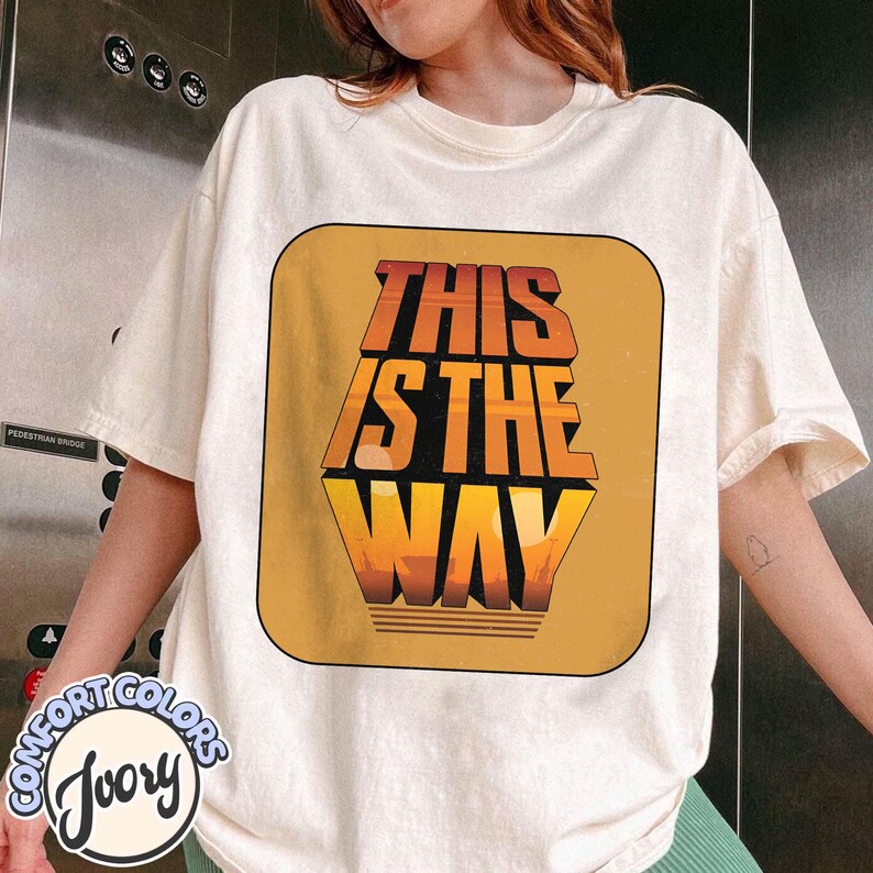 K&ouml;nnte beinhalten: Elfenbeinfarbenes T-Shirt mit einem Grafikdesign im Retro-Stil mit der Aufschrift "THIS IS THE WAY" in Orange und Gelb. Die Grafik befindet sich in einem rechteckigen Rahmen. Das Shirt ist aus Comfort Colors Material.