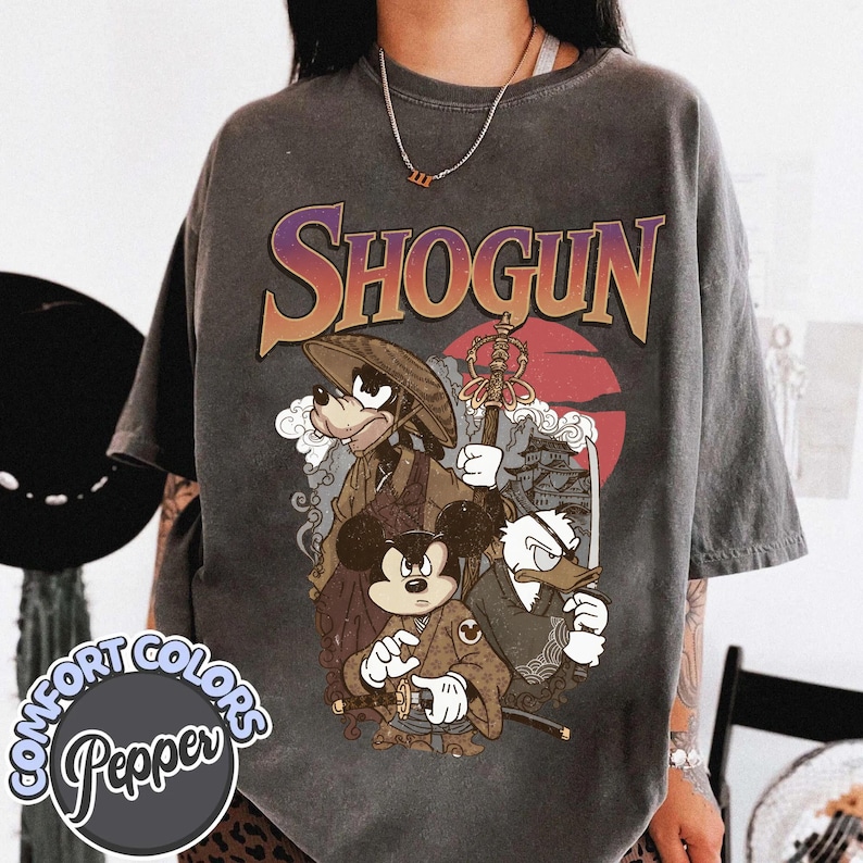 Puede incluir: Una camiseta de manga corta gris carb&oacute;n con un estampado gr&aacute;fico que incluye el texto SHOGUN en una fuente degradada. El dise&ntilde;o muestra tres personajes de dibujos animados vestidos con atuendos japoneses tradicionales, incluyendo un sombrero de samur&aacute;i, una espada y un bast&oacute;n, sobre un gran sol rojo y la silueta de un castillo.