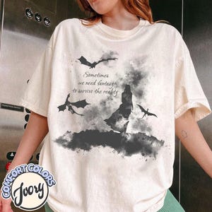 May include: An ivory short-sleeve t-shirt featuring a black watercolor-style graphic of a person standing amidst flying dragons and smoke. The text reads, Sometimes we need fantasy to survive the reality. The design is centered on the front of the shirt.