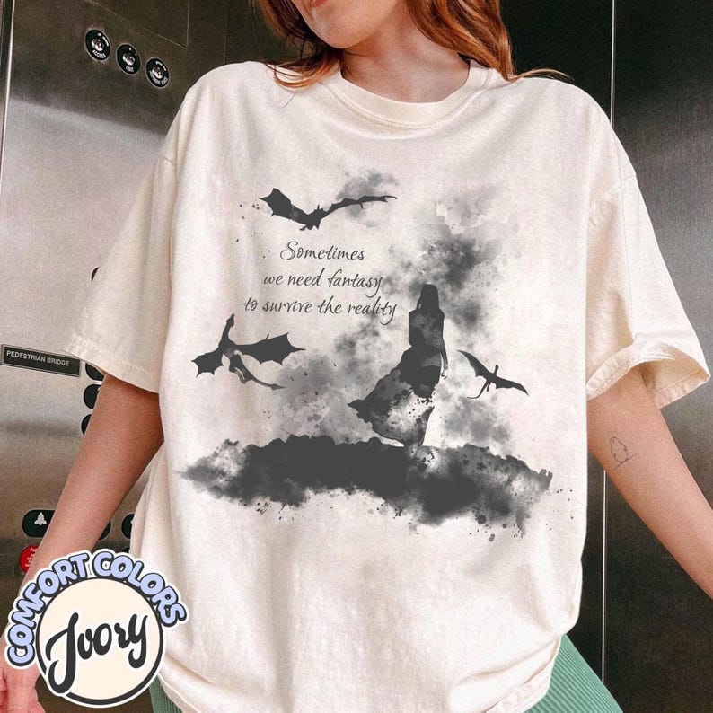 May include: An ivory short-sleeve t-shirt featuring a black watercolor-style graphic of a person standing amidst flying dragons and smoke. The text reads, Sometimes we need fantasy to survive the reality. The design is centered on the front of the shirt.
