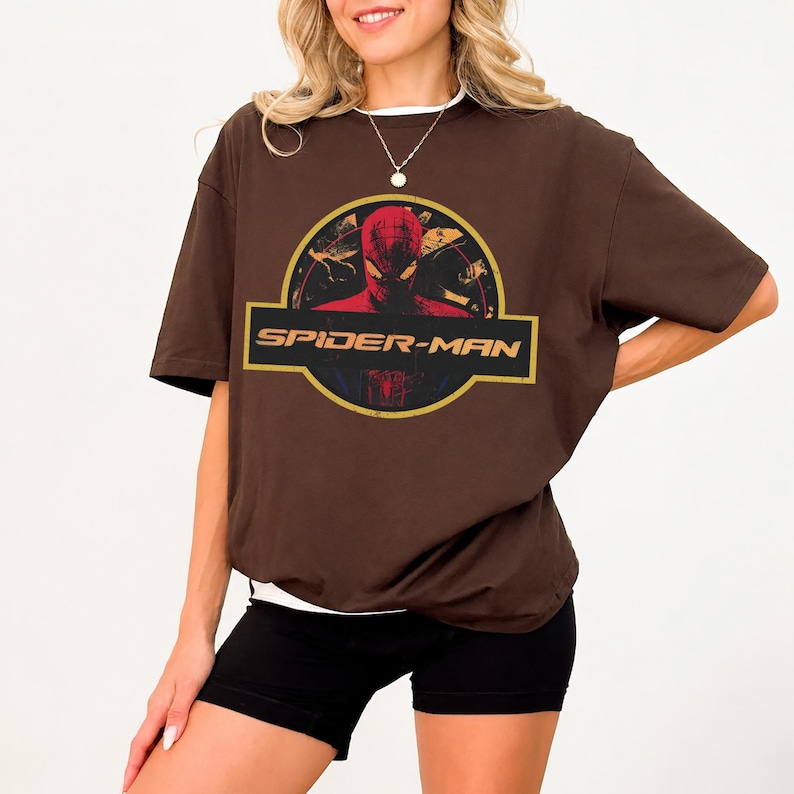 May include: A brown short-sleeved t-shirt featuring a graphic print of the Spider-Man character inside a circular yellow and black emblem. The text SPIDER-MAN is printed in a bold yellow font across a black horizontal bar within the design.