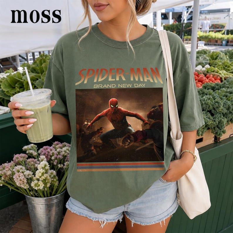 May include: An olive green short-sleeve t-shirt featuring a graphic print of Spider-Man in a dynamic action pose. The text "SPIDER-MAN" and "BRAND NEW DAY" appear above the image, with horizontal stripes in red, blue, and yellow below the graphic.