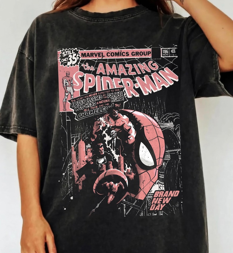 Peut inclure: T-shirt gris fonc&eacute; avec un design de couverture de bande dessin&eacute;e. Le design comprend le texte "The Amazing Spider-Man" en rose et blanc, des d&eacute;tails de bande dessin&eacute;e et une illustration de Spider-Man. Le t-shirt a un aspect vintage et us&eacute;.