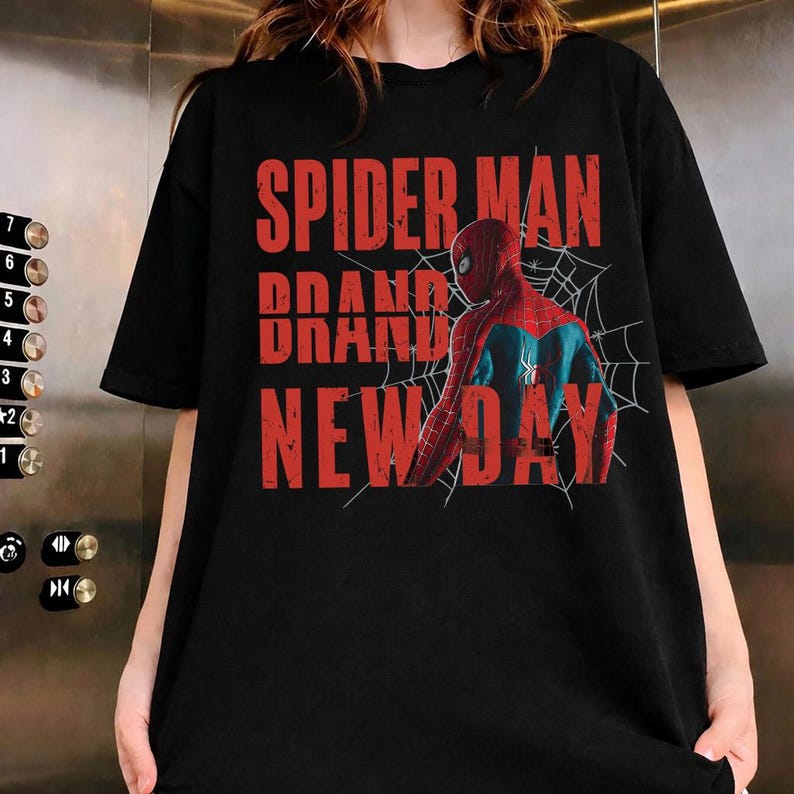 K&ouml;nnte beinhalten: Schwarzes T-Shirt mit dem roten Schriftzug "SPIDER MAN BRAND NEW DAY". Das Design zeigt eine Grafik von Spider-Man in seinem rot-blauen Anzug mit einem Spinnennetz. Das T-Shirt ist ein l&auml;ssiger Stil.
