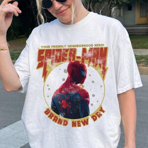 May include: White t-shirt featuring a graphic of Spider-Man in a red and blue suit, with the text "YOUR FRIENDLY NEIGHBORHOOD HERO! SPIDER-MAN BRAND NEW DAY" in red and yellow. The design is vintage-inspired.